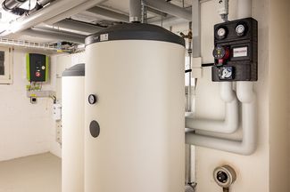 Local technique avec deux grands chauffe-eau cylindriques à accumulation ainsi que différentes conduites, ar-matures et module de commande monté au mur.